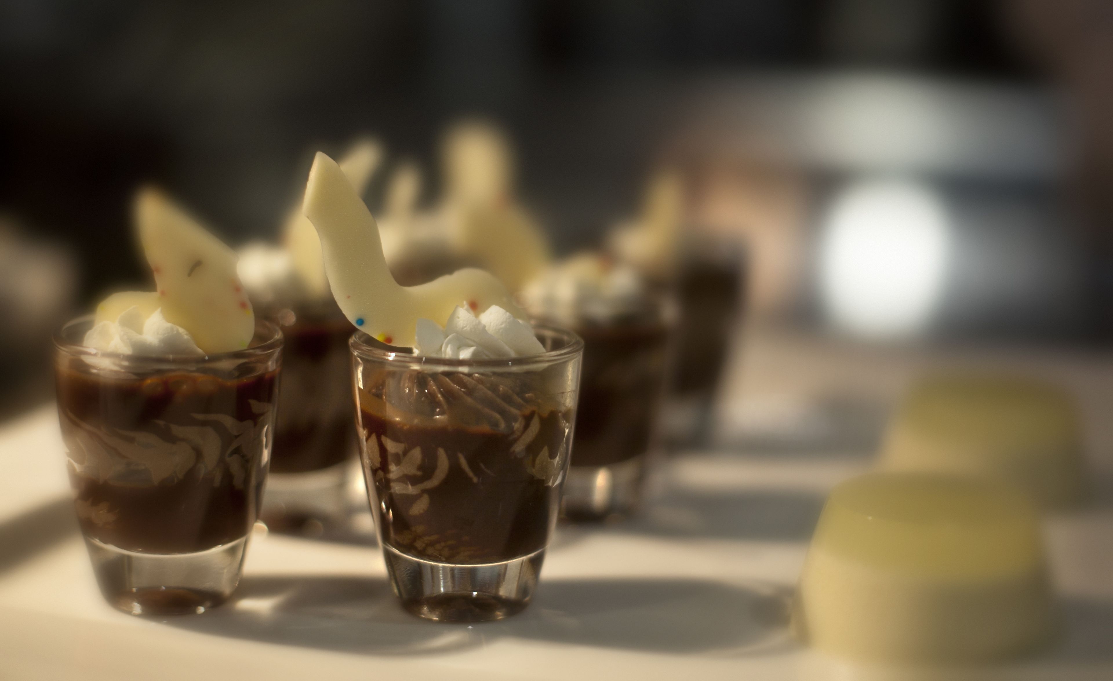 A series of small glasses containing a dark liquid, possibly a dessert or drink, with whipped cream and a slice of fruit on top.