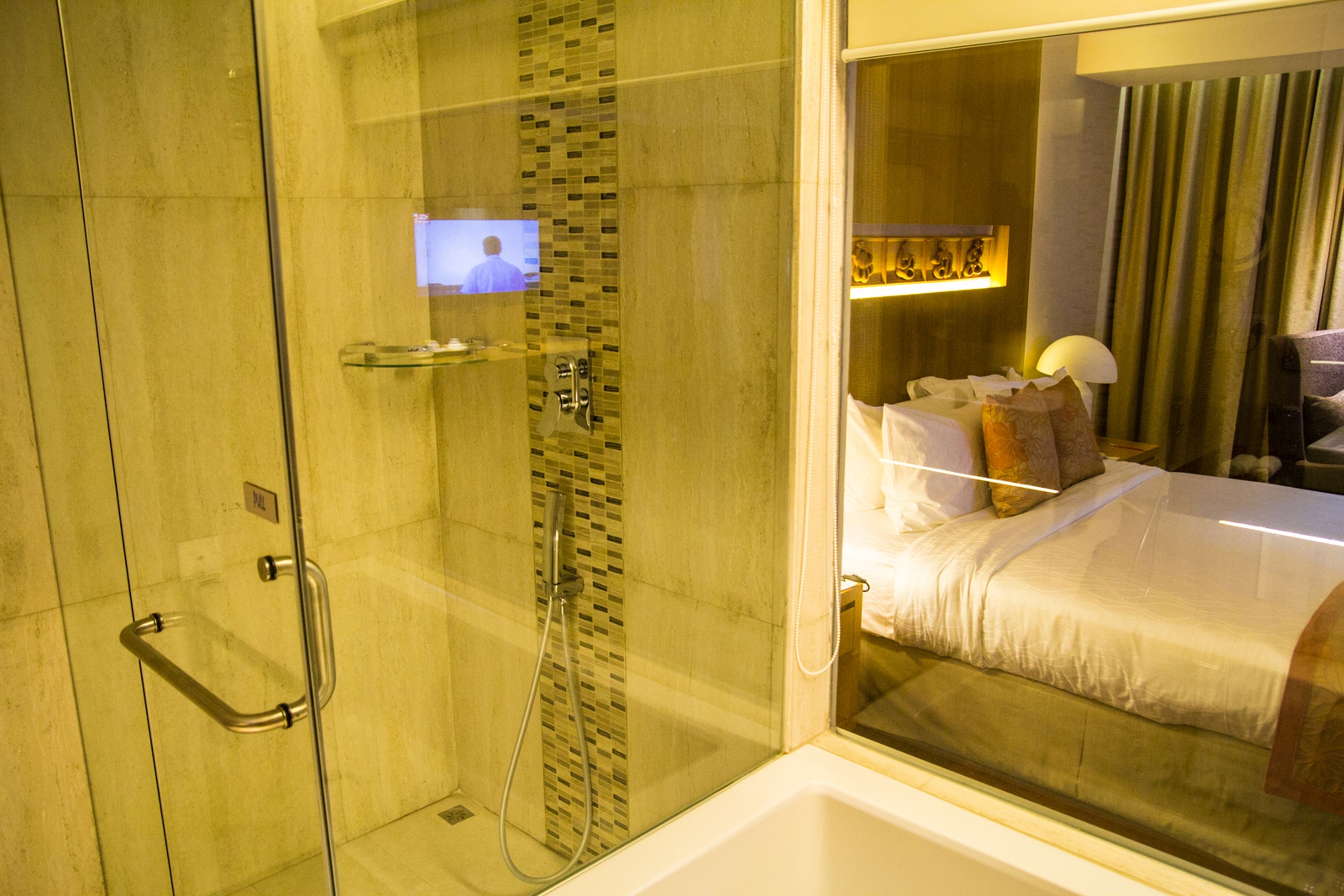 Quarto de hotel com box de vidro, cama com roupa de cama branca e televisão visível.