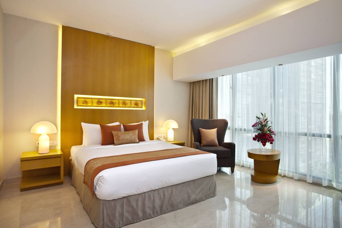 A modern hotel room with a large bed, bedside lamps, a chair, a side table, and a large window with curtains.