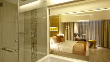 A modern hotel room with a glass shower enclosure, a large bed with multiple pillows, a bedside table, and a view of the room through a glass partition.
