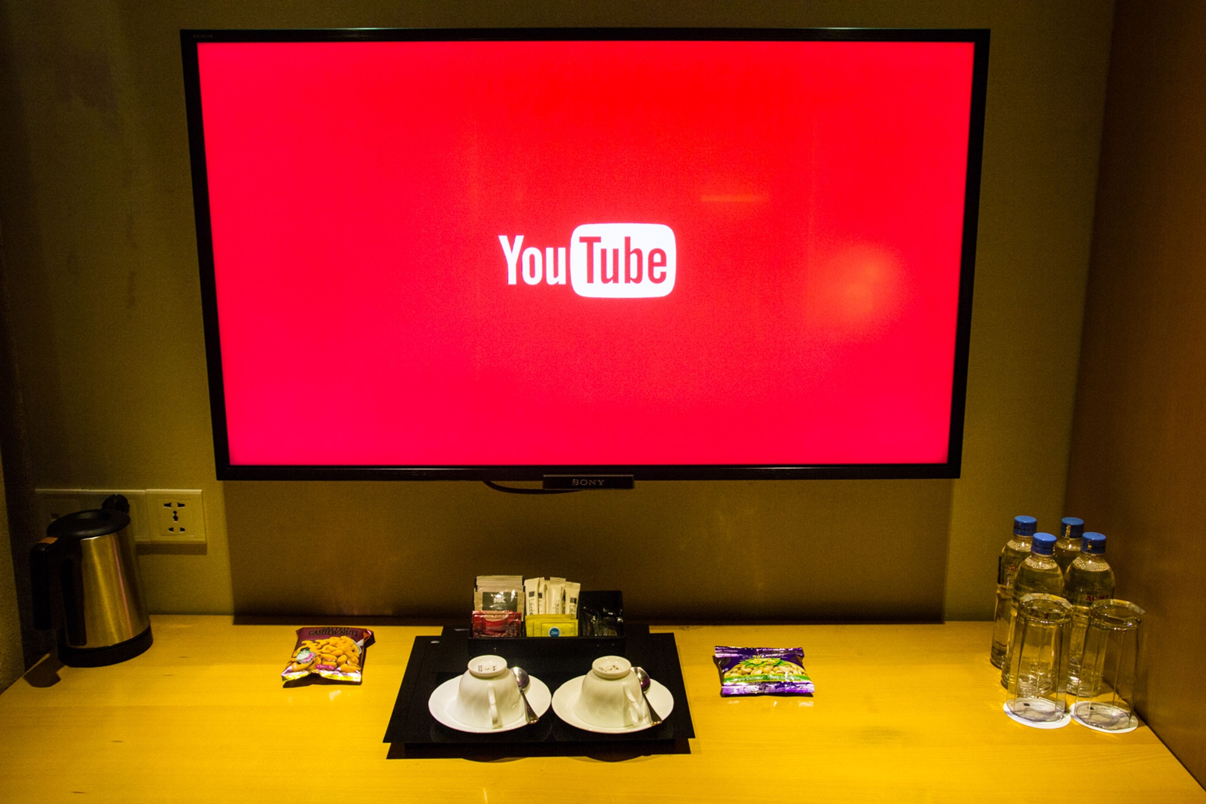 Un televisor mostrando el logotipo de YouTube, una cafetera, un juego de té, bocadillos y agua embotellada sobre una mesa.