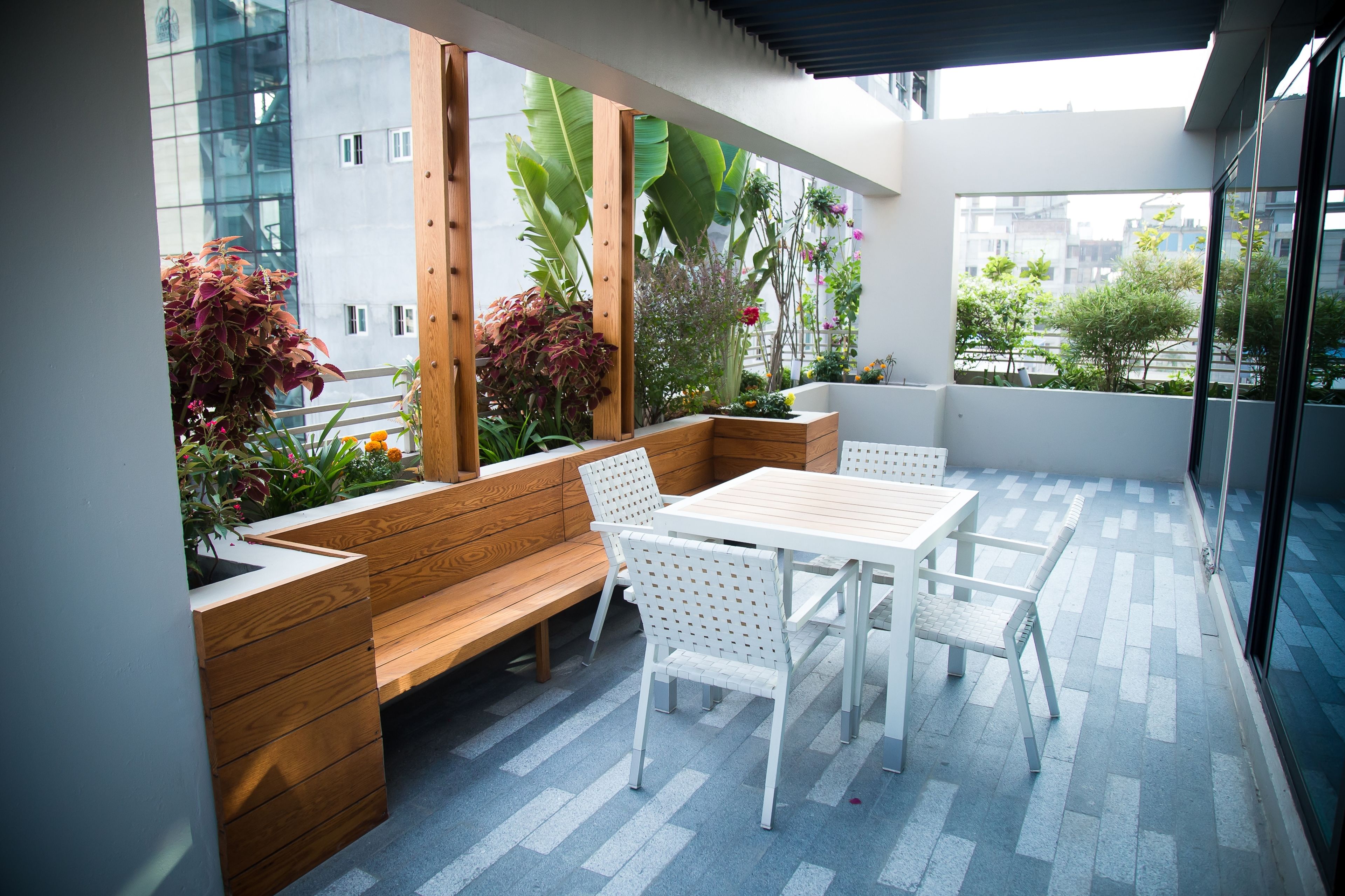 A modern outdoor seating area with a wooden bench and a table, surrounded by potted plants and a view of the cityscape.
