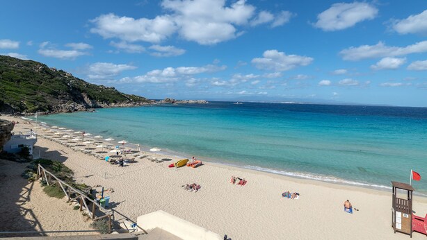 Een strand met helder turquoise water, een houten redershut en strandparasols.