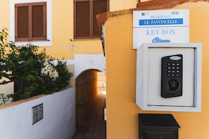 A key box with a digital keypad, a sign indicating 'Residence Le Pavoncelle Key Box', and a yellow building with brown shutters in the background.