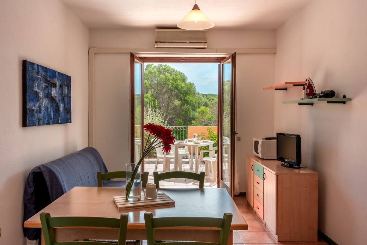 Un coin repas comprenant une table, des chaises, un vase avec des fleurs, un banc, un téléviseur posé sur un meuble et une vue sur la verdure à l’extérieur.