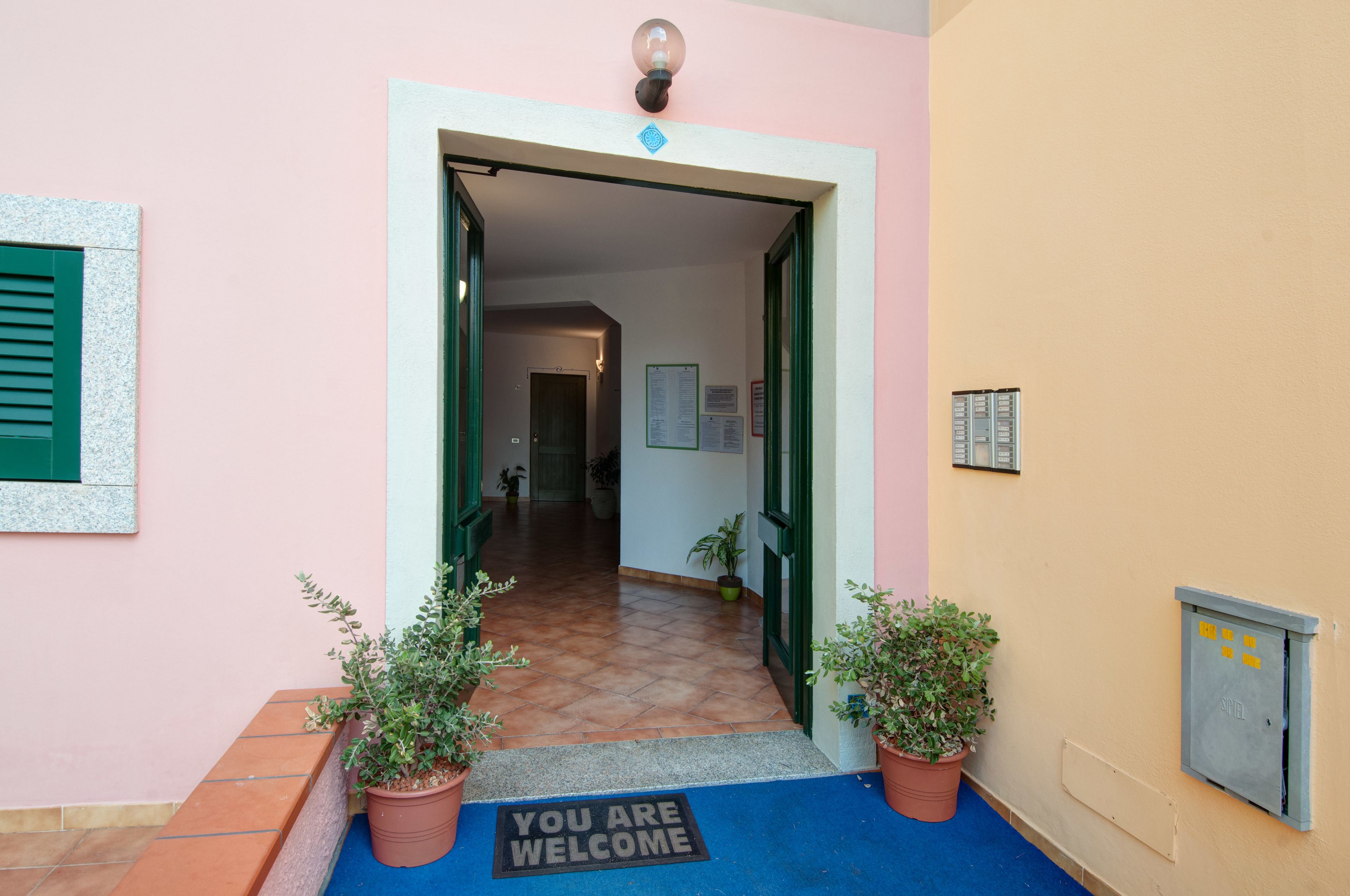 A hotel entrance with a 'You are welcome' mat, potted plants, and a blue carpet leading to the door.