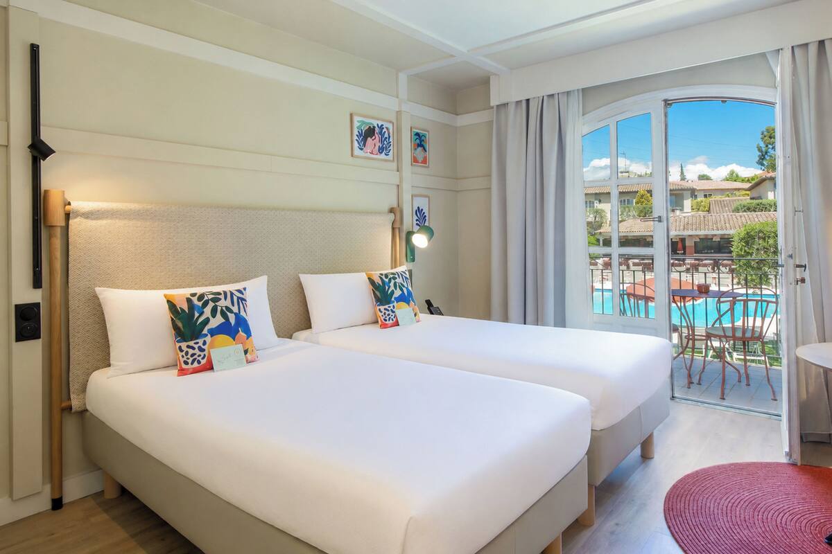 Habitación de hotel con dos camas, un balcón con vistas a una piscina y edificios, y una alfombra roja.