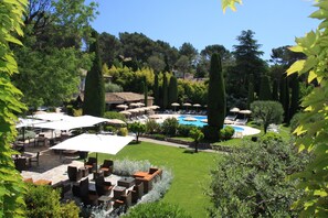 A poolside area with outdoor seating and umbrellas, surrounded by lush greenery and tall trees.