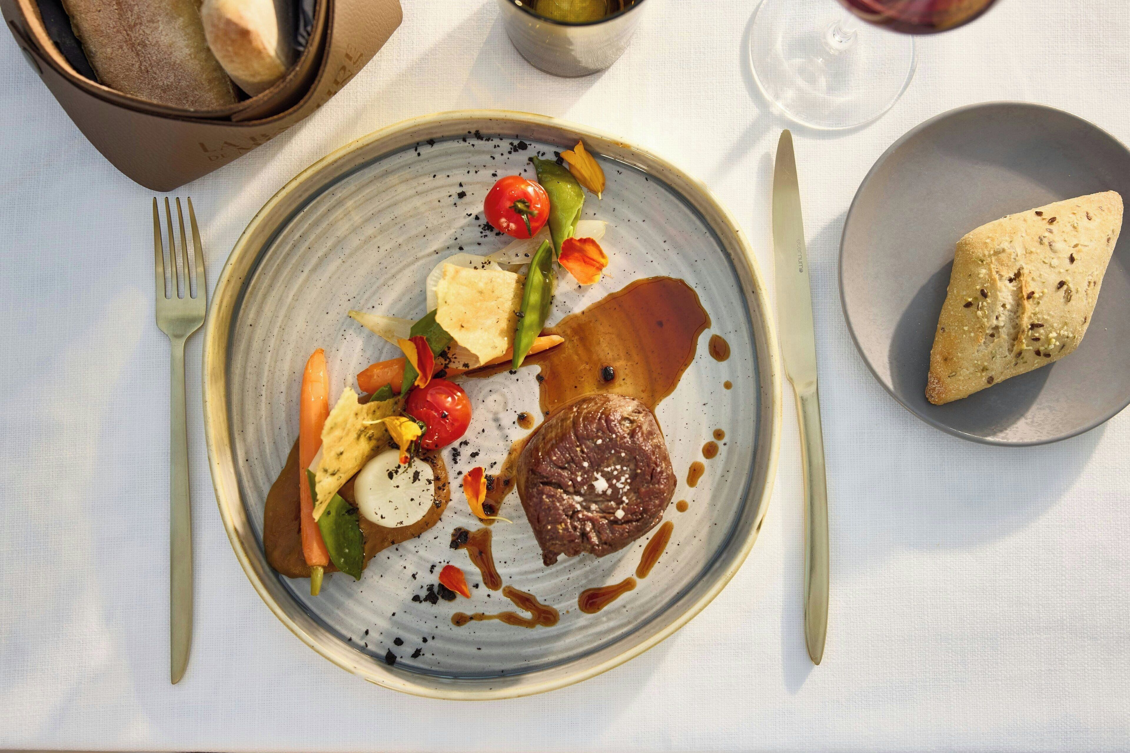 Un plato con carne, verduras y salsa, un tenedor, cuchillo y un bol de pan sobre una mesa.