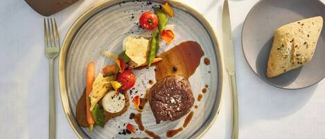Un plato con carne, verduras y salsa, un tenedor, cuchillo y un bol de pan sobre una mesa.