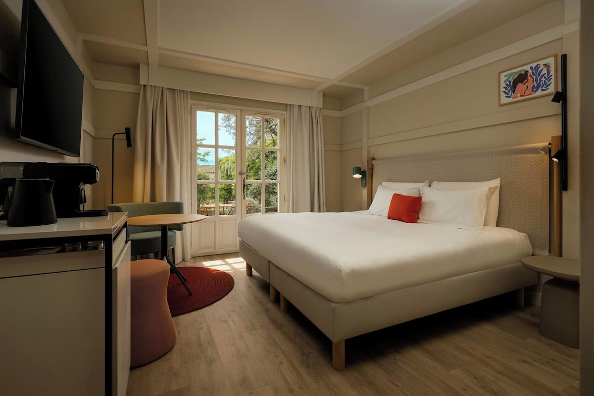 Habitación de hotel con cama, escritorio con cafetera, televisor y ventana con cortinas.