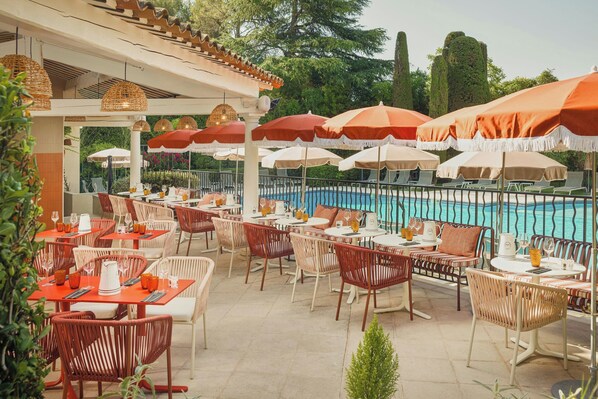 An outdoor dining area with tables and chairs, umbrellas, and a pool in the background.