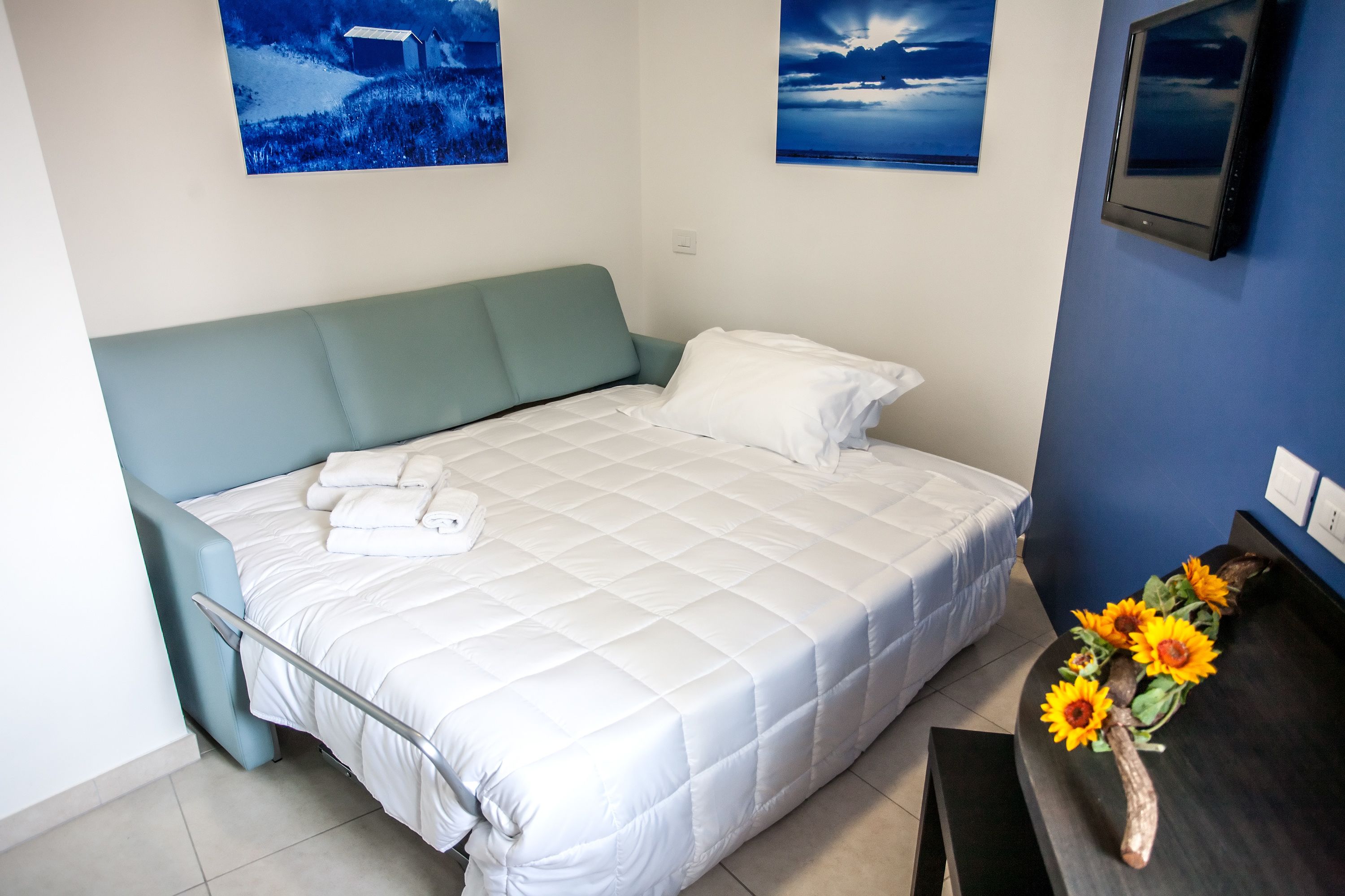 Una cama bien hecha con ropa de cama blanca, un cabecero color aguamarina y una mesita de noche con un jarrón de girasoles.