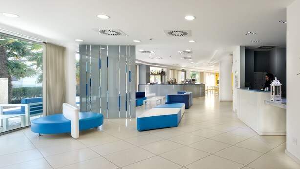 Un moderno vestíbulo de hotel con zonas de estar, mostrador de recepción y amplios ventanales con vistas a palmeras.