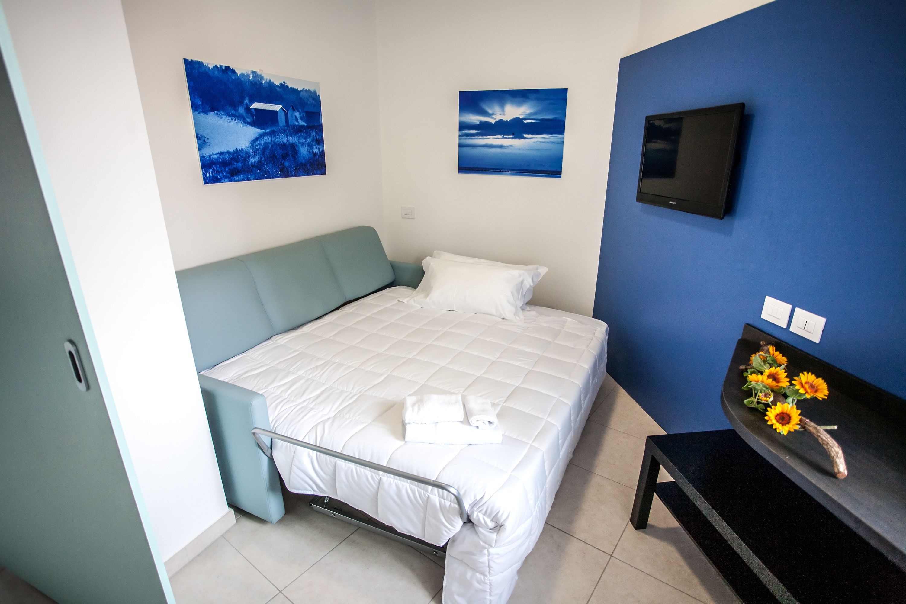 A neatly made bed with a white comforter and pillows, a flat-screen TV on a blue wall, and a small table with a vase of sunflowers.