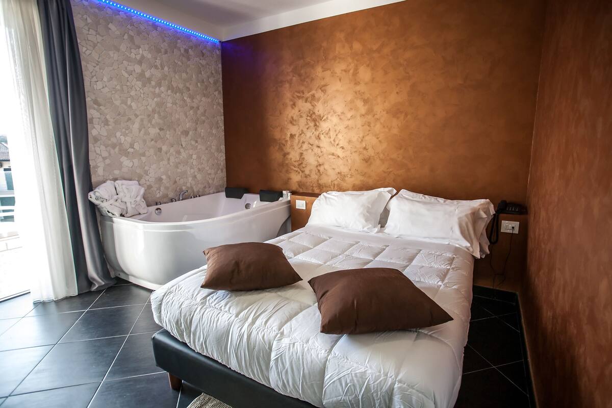 A modern hotel room with a large bed, a separate bathtub, and a stone-textured wall.