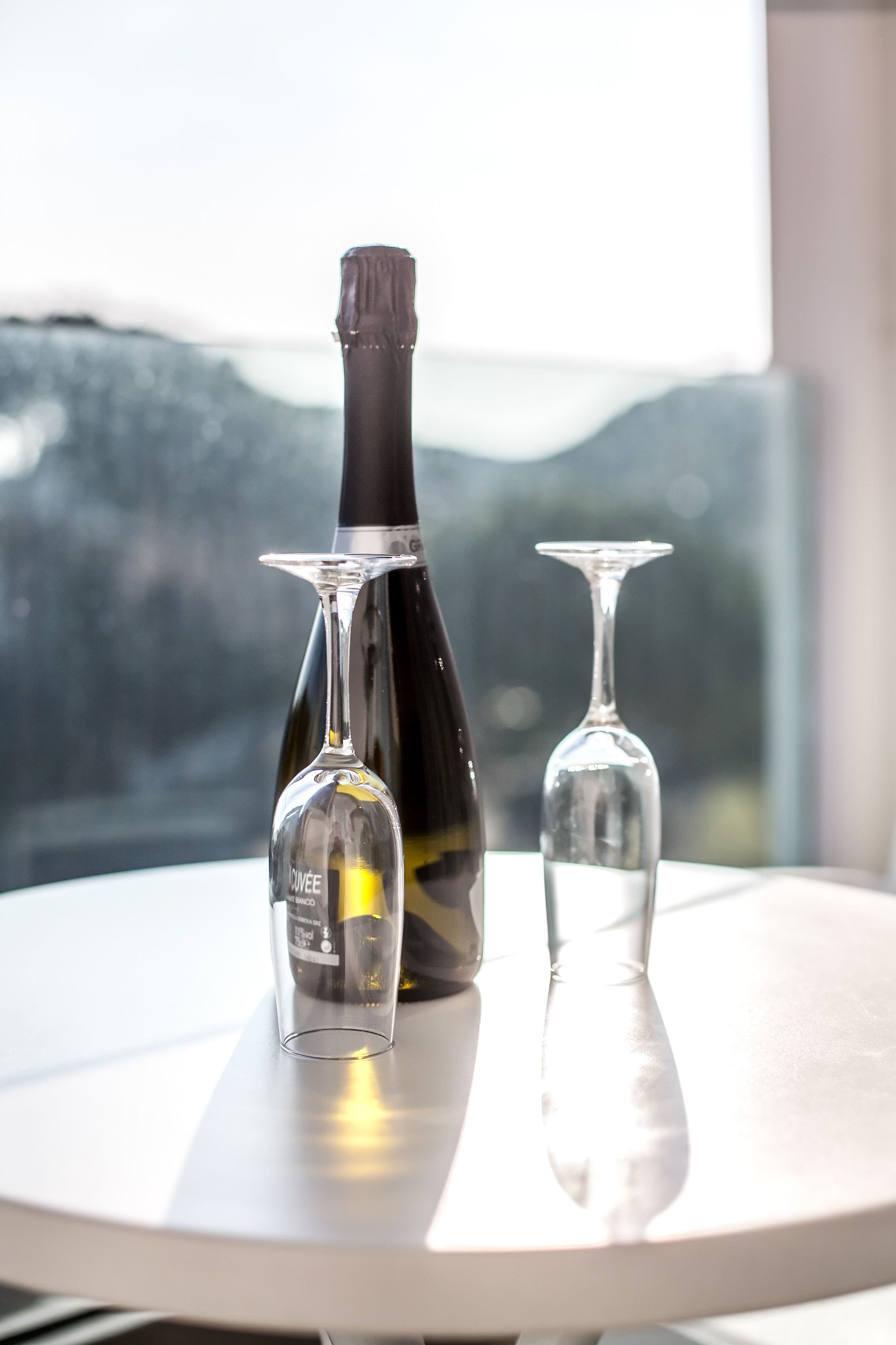 A bottle and two wine glasses on a table with a view of a mountainous landscape.