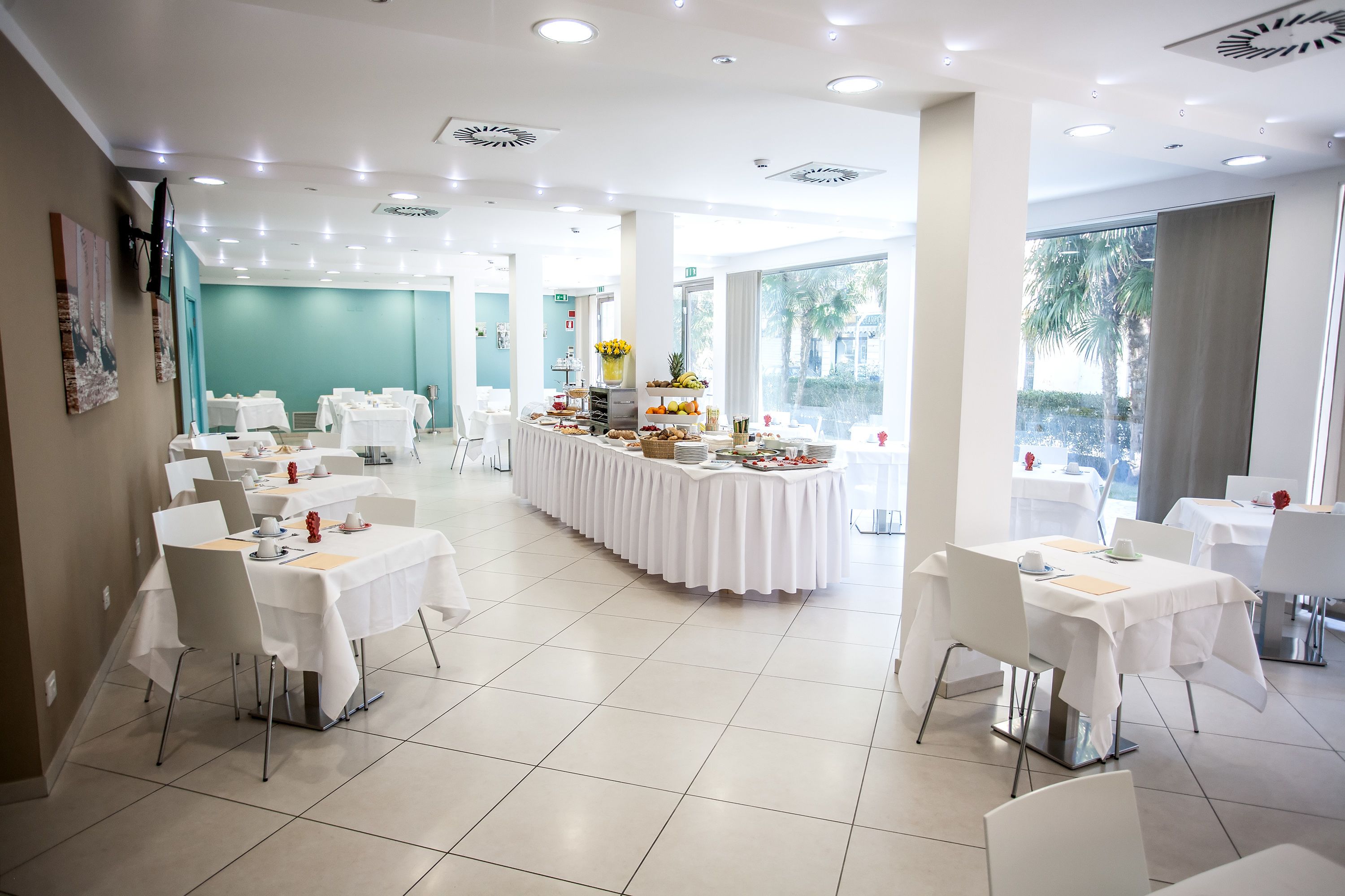 A spacious dining area with multiple tables and chairs, a central buffet with various food items, and large windows providing natural light.