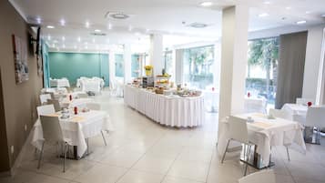 A spacious dining area with multiple tables and chairs, a central buffet with various food items, and large windows providing natural light.
