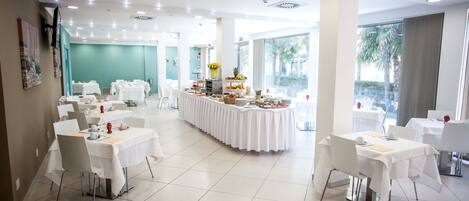 A spacious dining area with multiple tables and chairs, a central buffet with various food items, and large windows providing natural light.