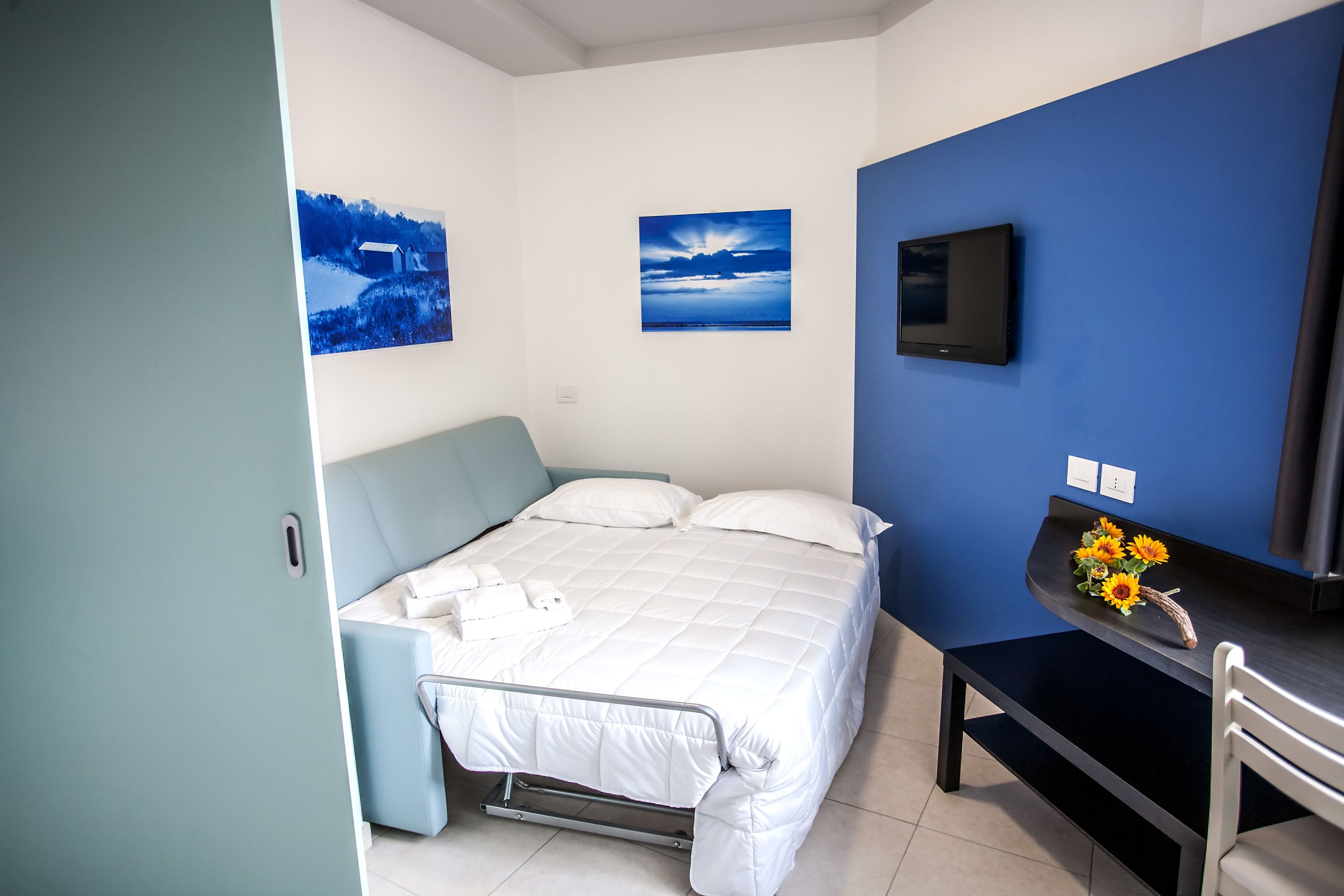 Un dormitorio con una cama, un escritorio con silla, un televisor y dos cuadros colgados en la pared.