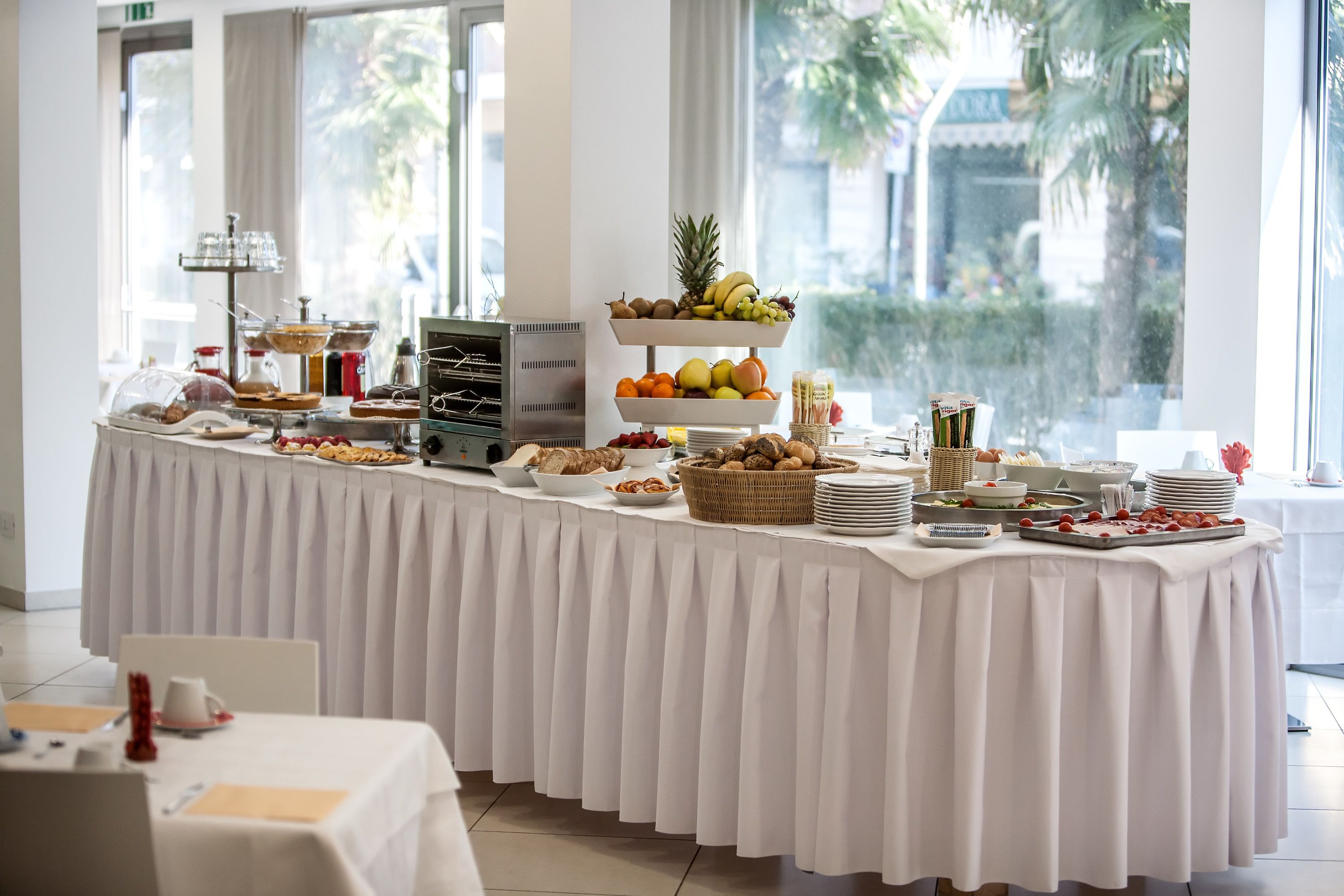 A buffet table with various food items and a fruit display, set in a bright room with large windows.