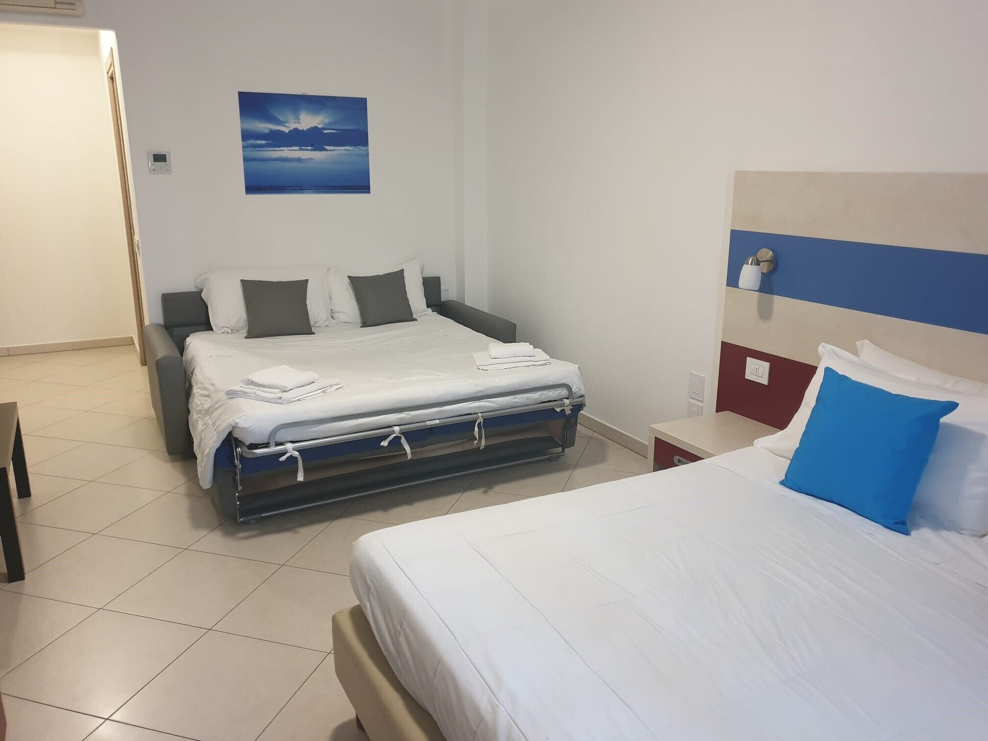 Et hotelværelse med en seng, et natbord, et vægur på væggen og et vindue med udsigt til himlen.