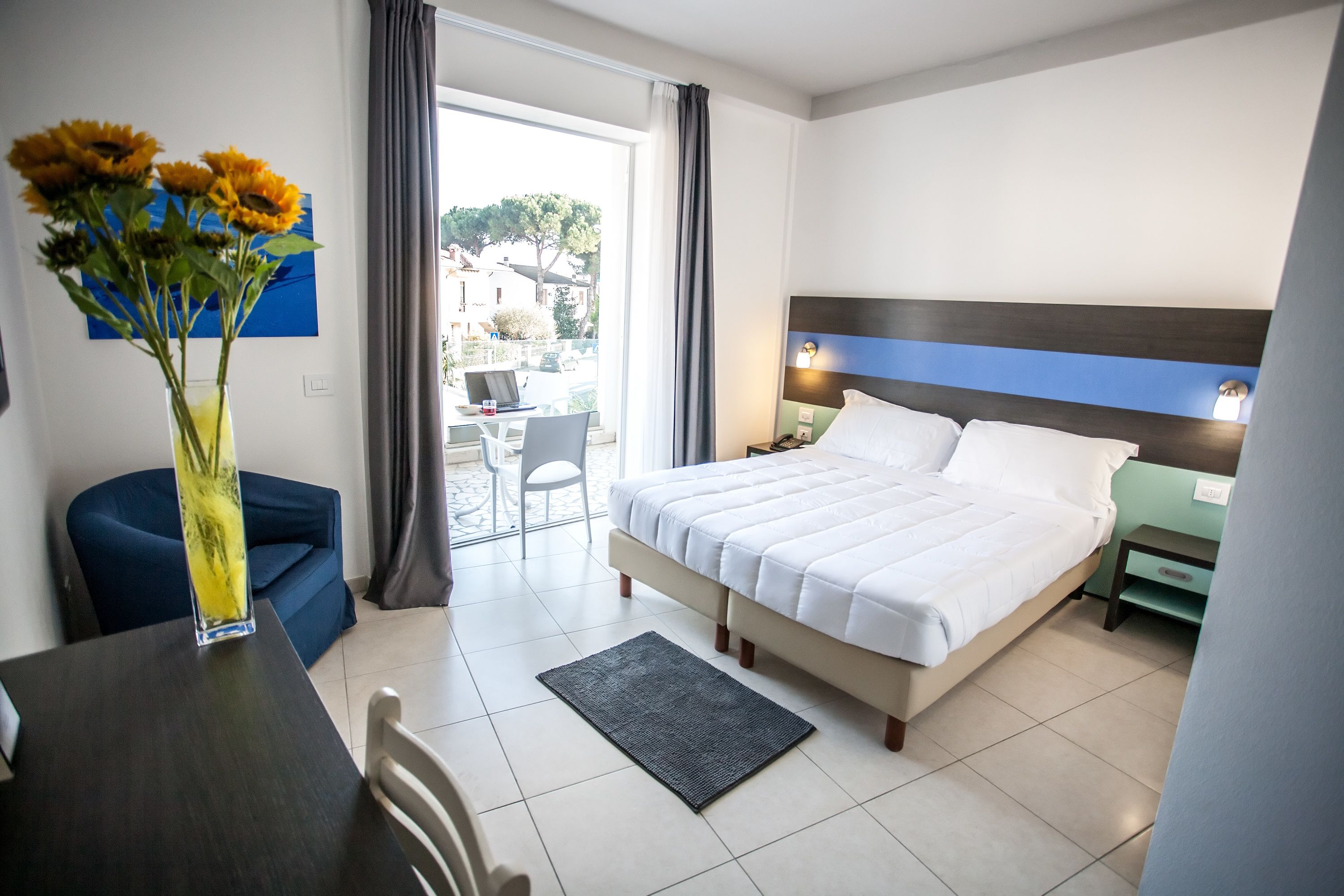 Ein Hotelzimmer mit einem Bett, einem Schreibtisch, einem Sessel, einer Vase mit Blumen und Blick ins Freie.