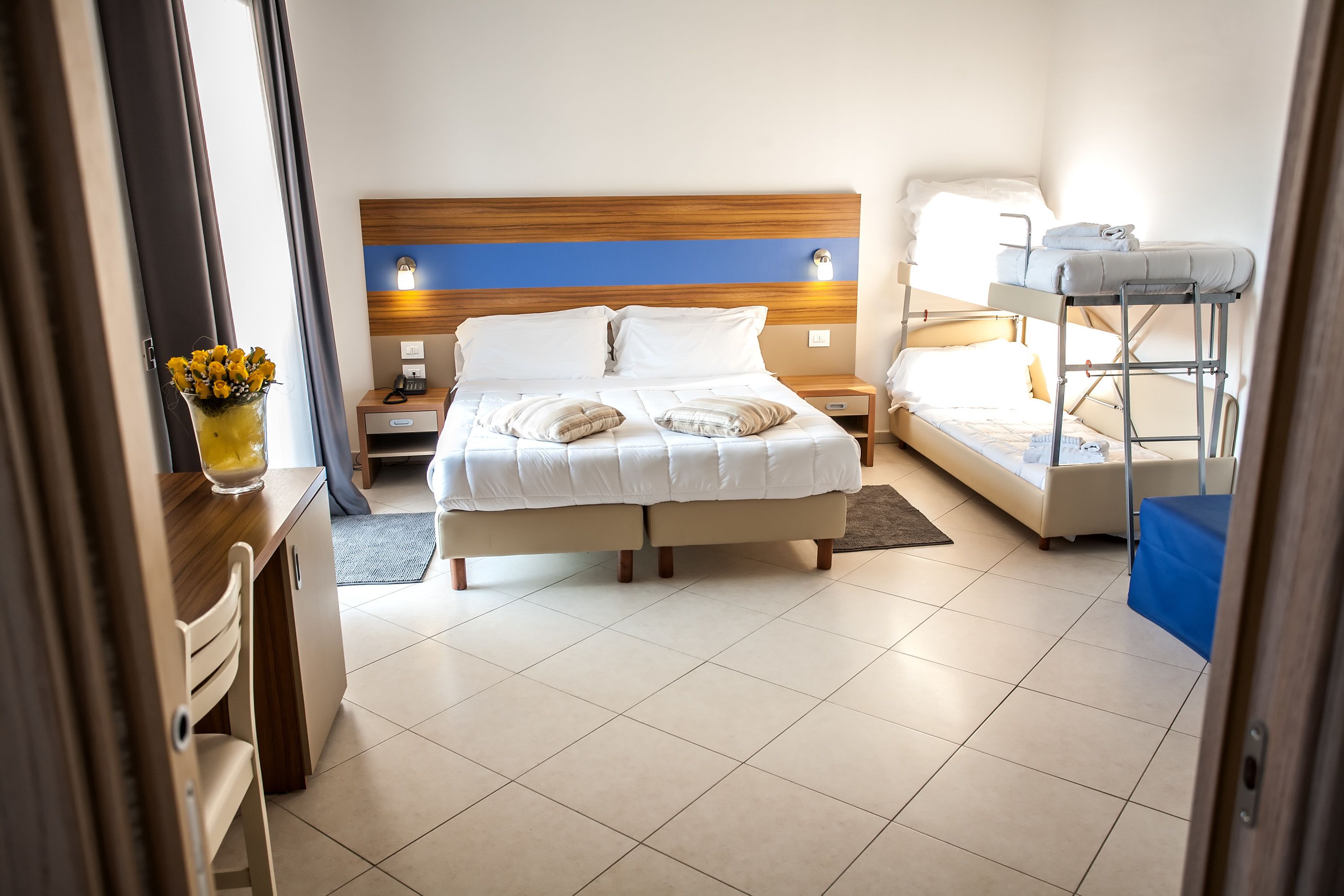 A hotel room with a bed, a desk, a chair, a bedside table, and a bunk bed.