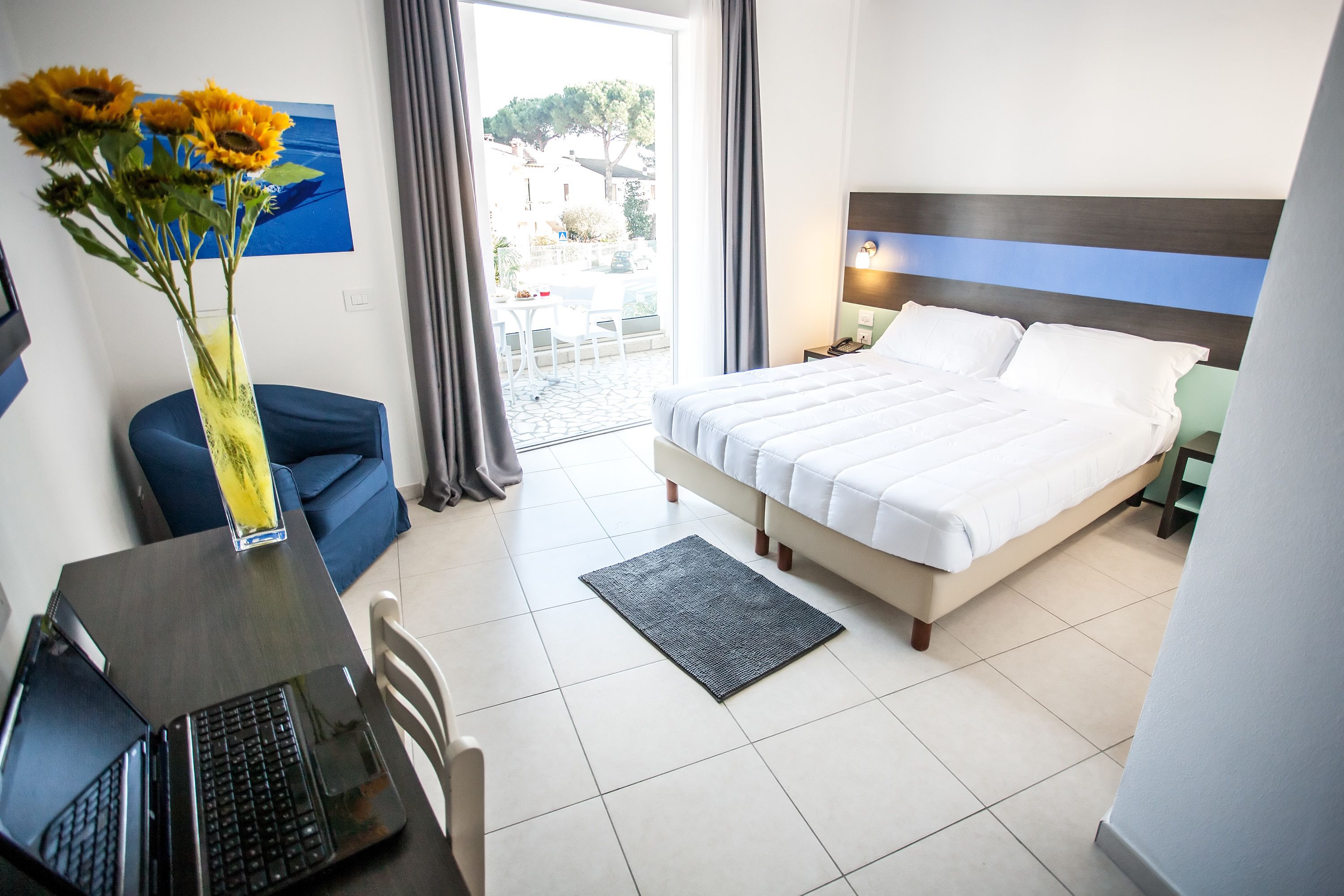 Quarto de hotel com cama, escrivaninha com laptop, um vaso de flores e vista para o exterior.