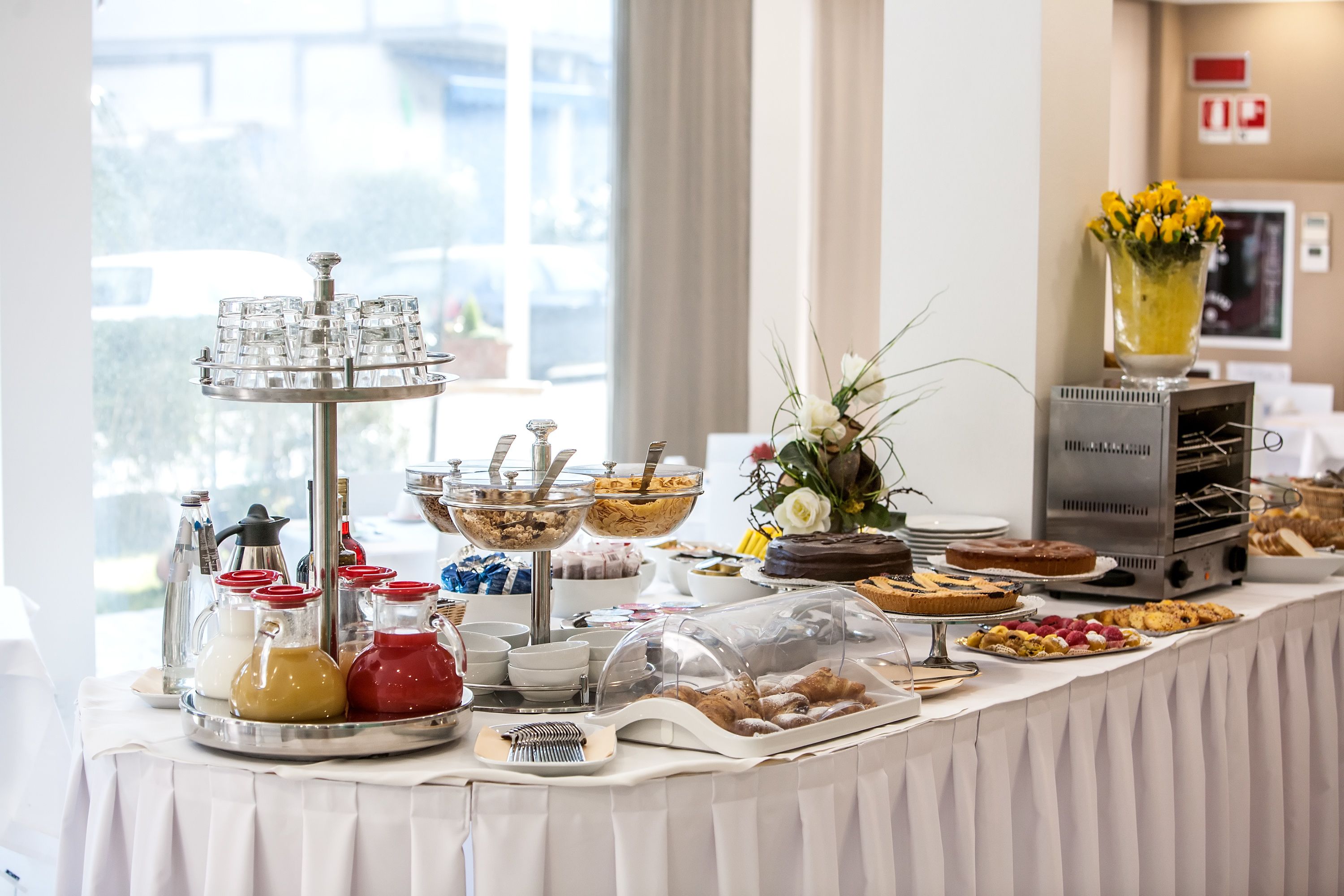 Una mesa de buffet con distintos alimentos y condimentos, una estación de café y un florero decorativo con flores.