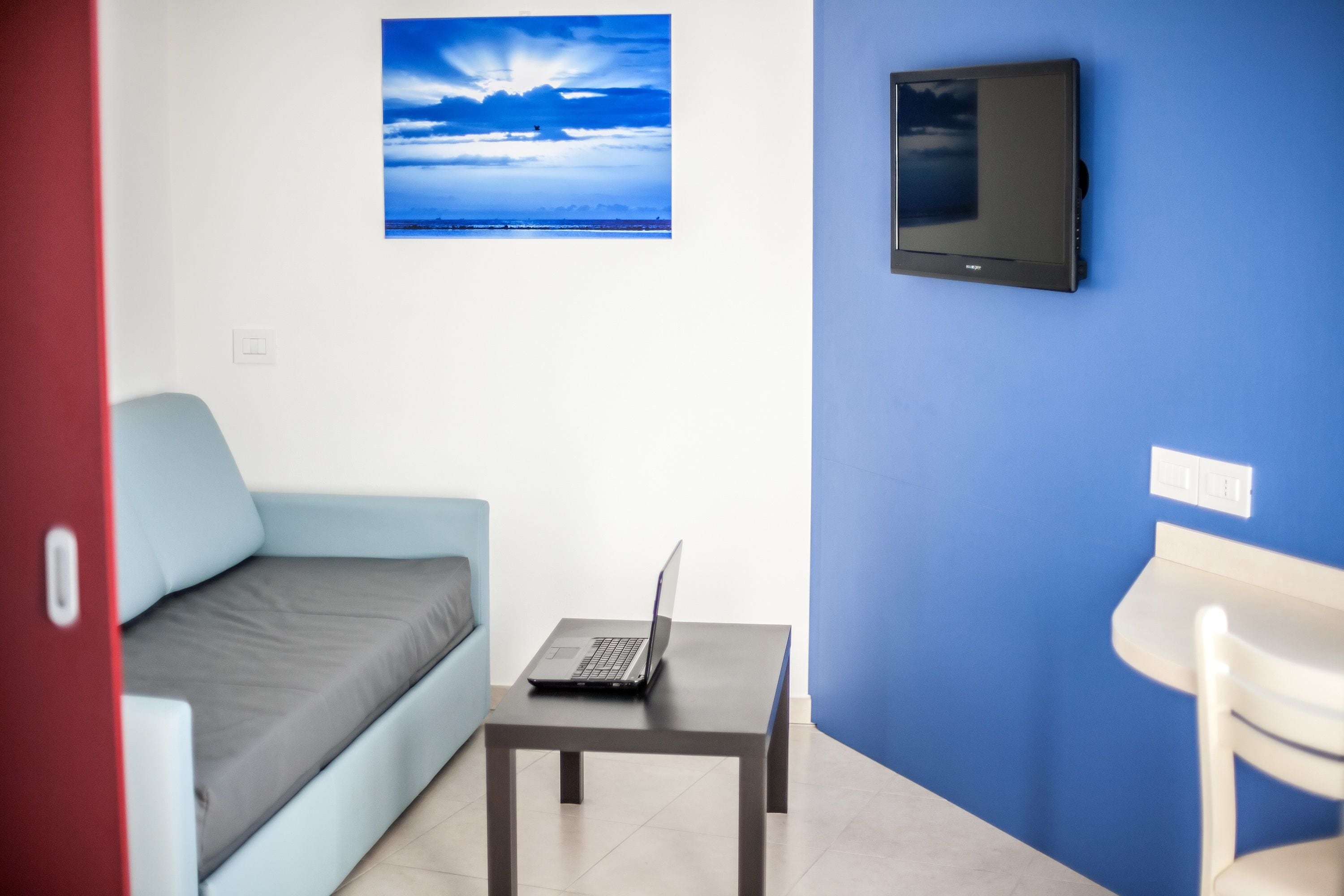 A room with a blue wall, a mounted TV, a small table with a laptop, a light blue sofa, and a white chair.