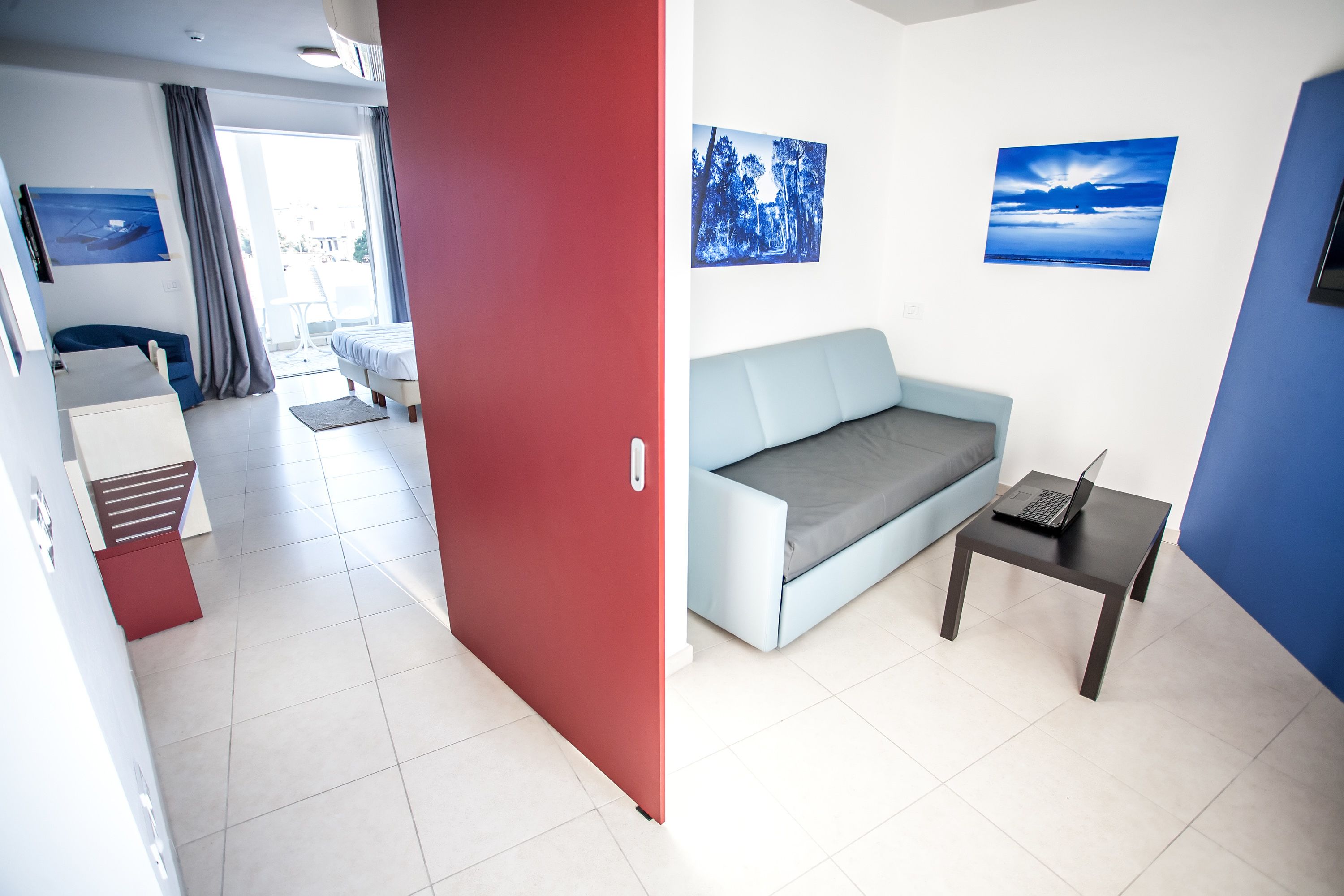 Ein modernes Hotelzimmer mit einer roten Tür, einem hellblauen Sofa, einem kleinen Tisch mit einem Laptop und zwei gerahmten Bildern an der Wand.