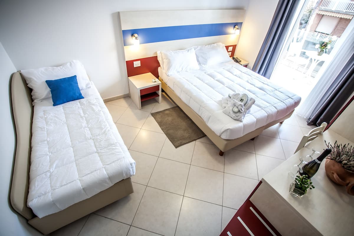 A hotel room with two beds, a small table with a bottle and glasses, and a view of the outside.