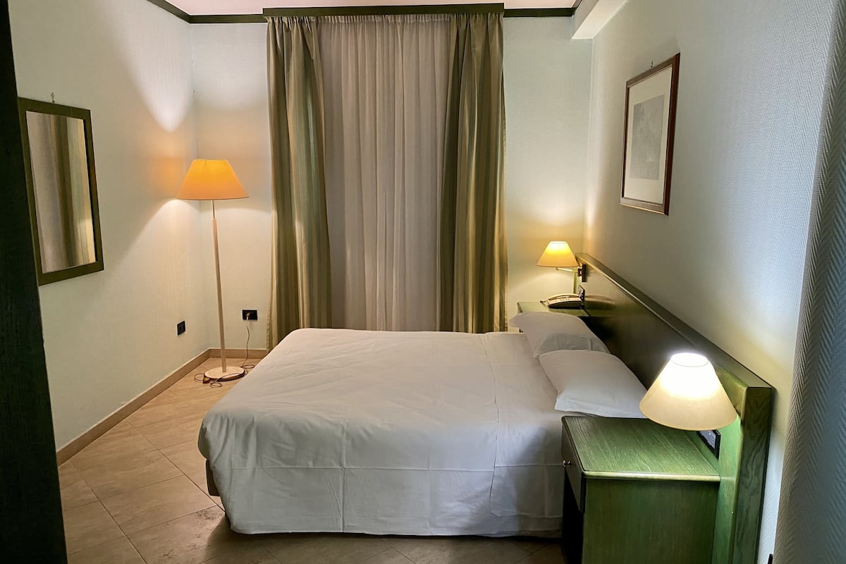 Quarto de hotel com uma cama, uma mesa de cabeceira com abajur, telefone fixado na parede e uma janela grande com cortinas.