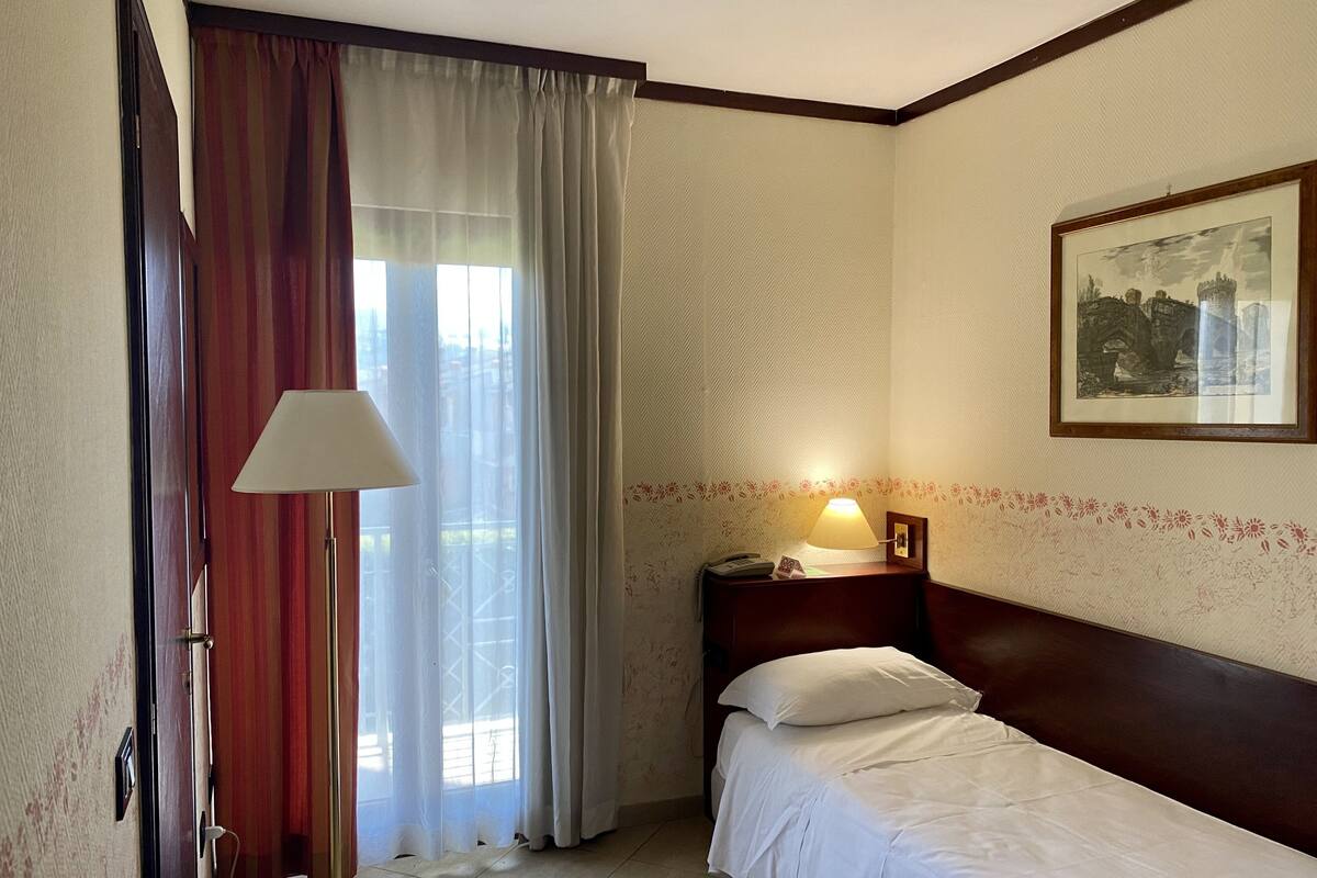 Quarto de hotel com uma cama de solteiro, mesa de cabeceira com abajur, janela com cortinas e um quadro na parede.