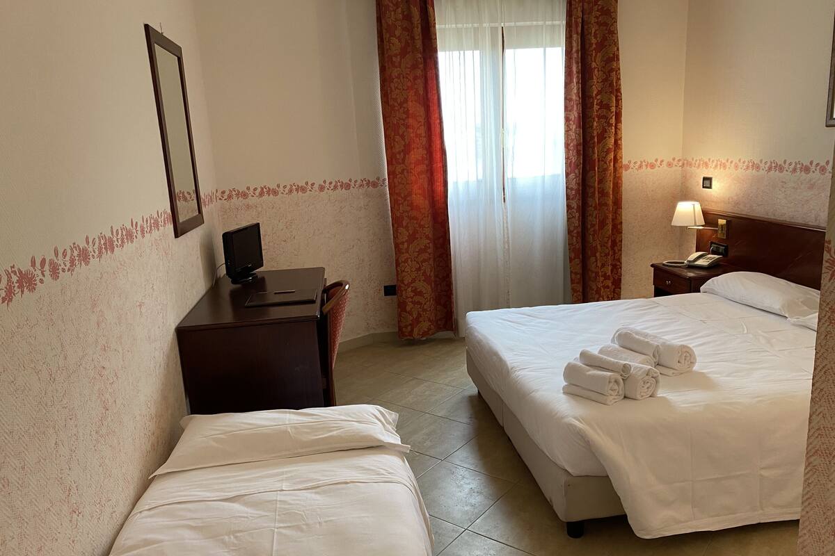 Quarto de hotel com duas camas, uma escrivaninha com computador, mesa de cabeceira com luminária e janela com cortinas.