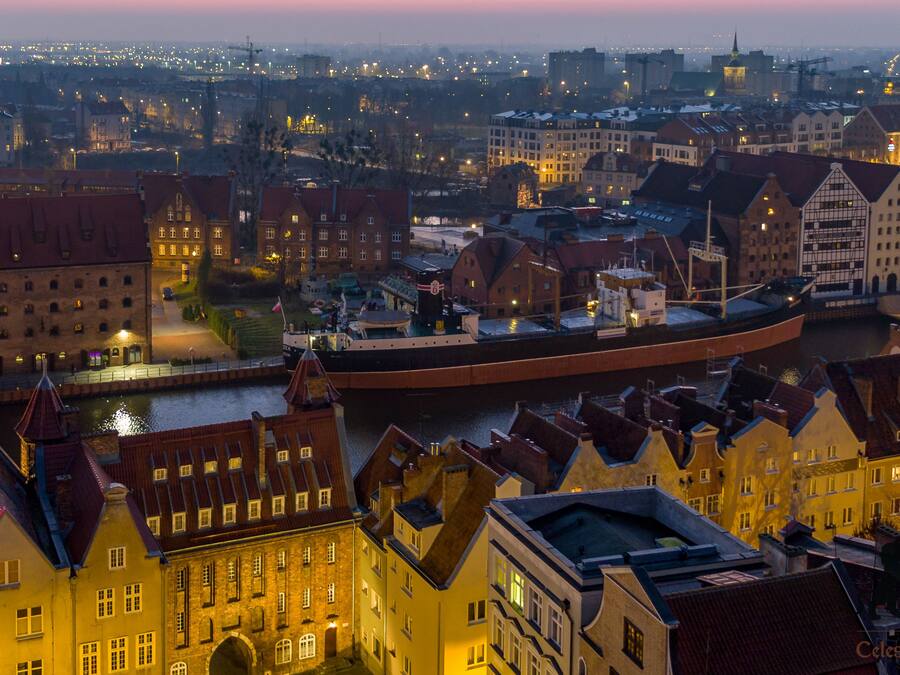A historic cityscape with traditional architecture, a river, and a large ship docked at the harbor.