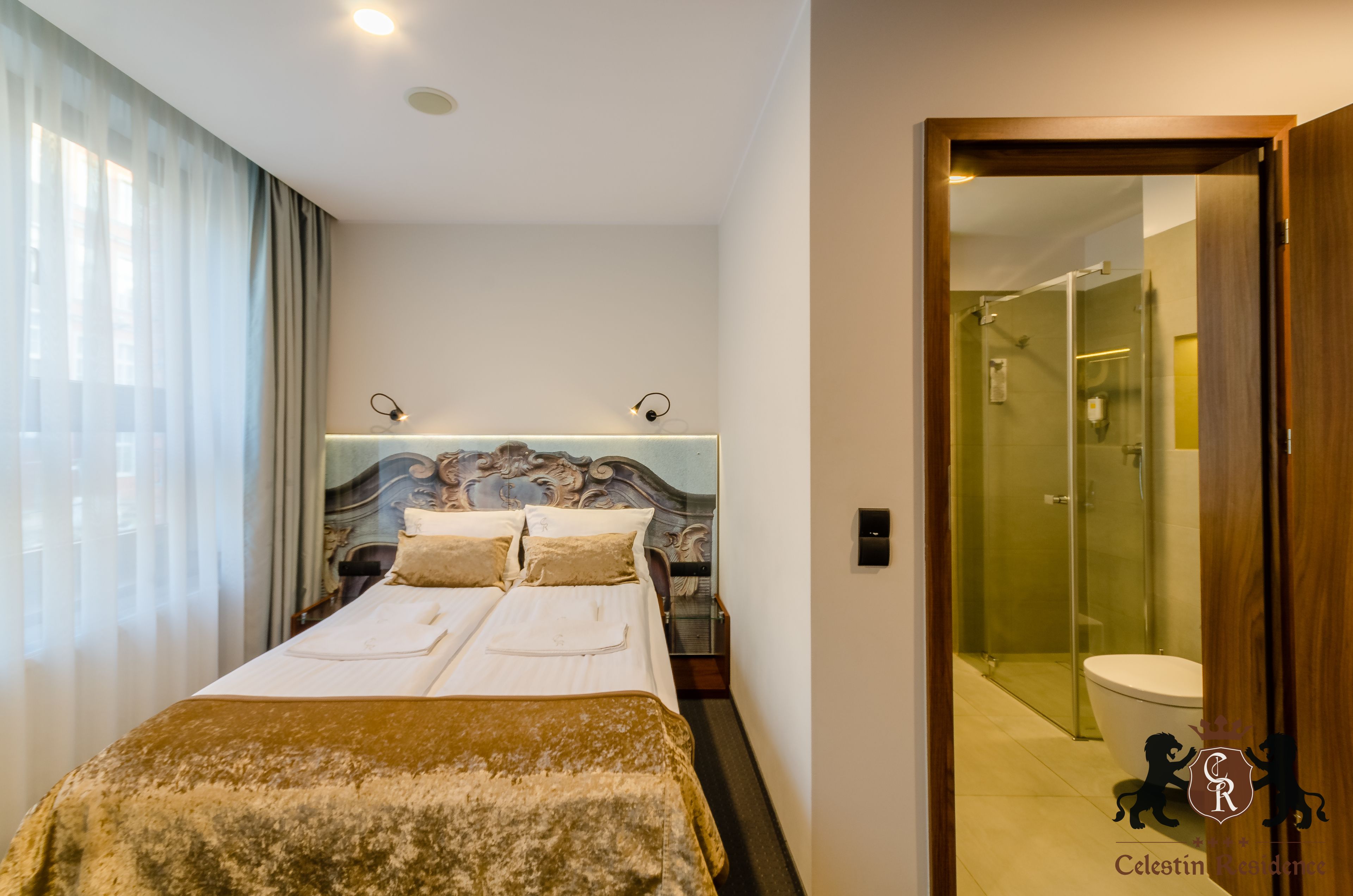 A hotel room with a double bed, a bedside table, a nightstand, a headboard with a decorative design, a window with curtains, and a bathroom visible through a glass door.