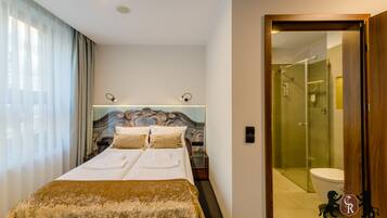 A hotel room with a double bed, a bedside table, a nightstand, a headboard with a decorative design, a window with curtains, and a bathroom visible through a glass door.