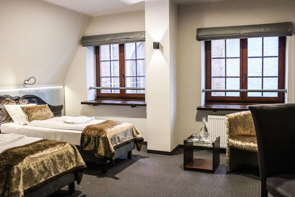 A hotel room with two beds, a chair, a small table, and windows with views of a brick building.