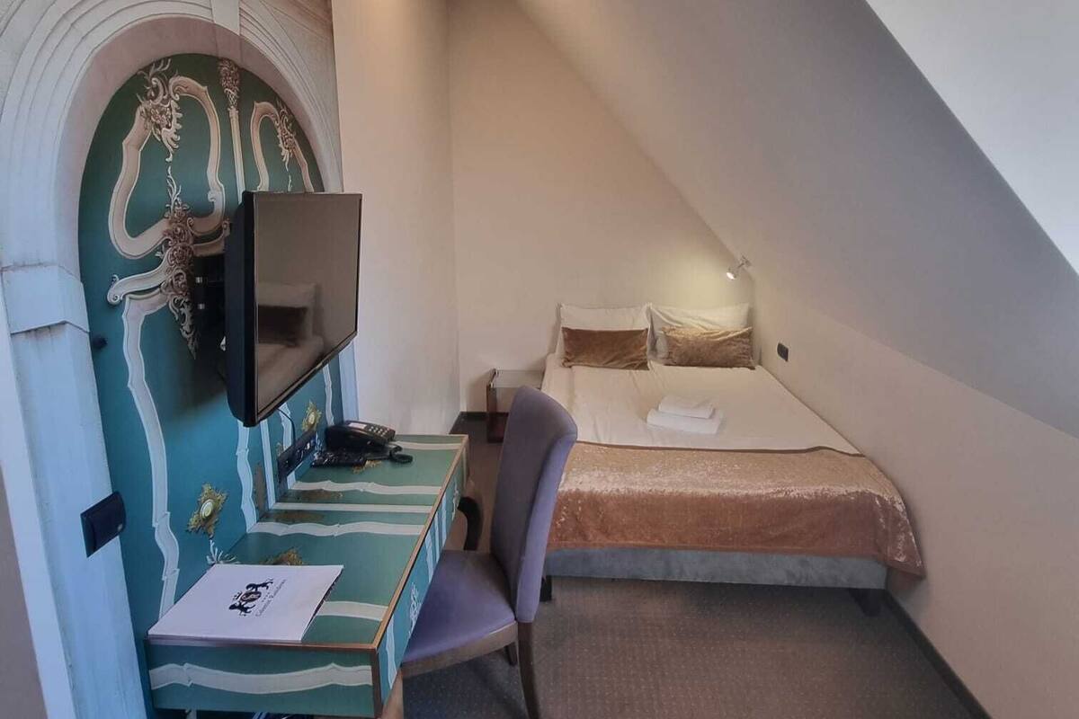 A small, triangular room with a bed, a desk with a television, and a chair.