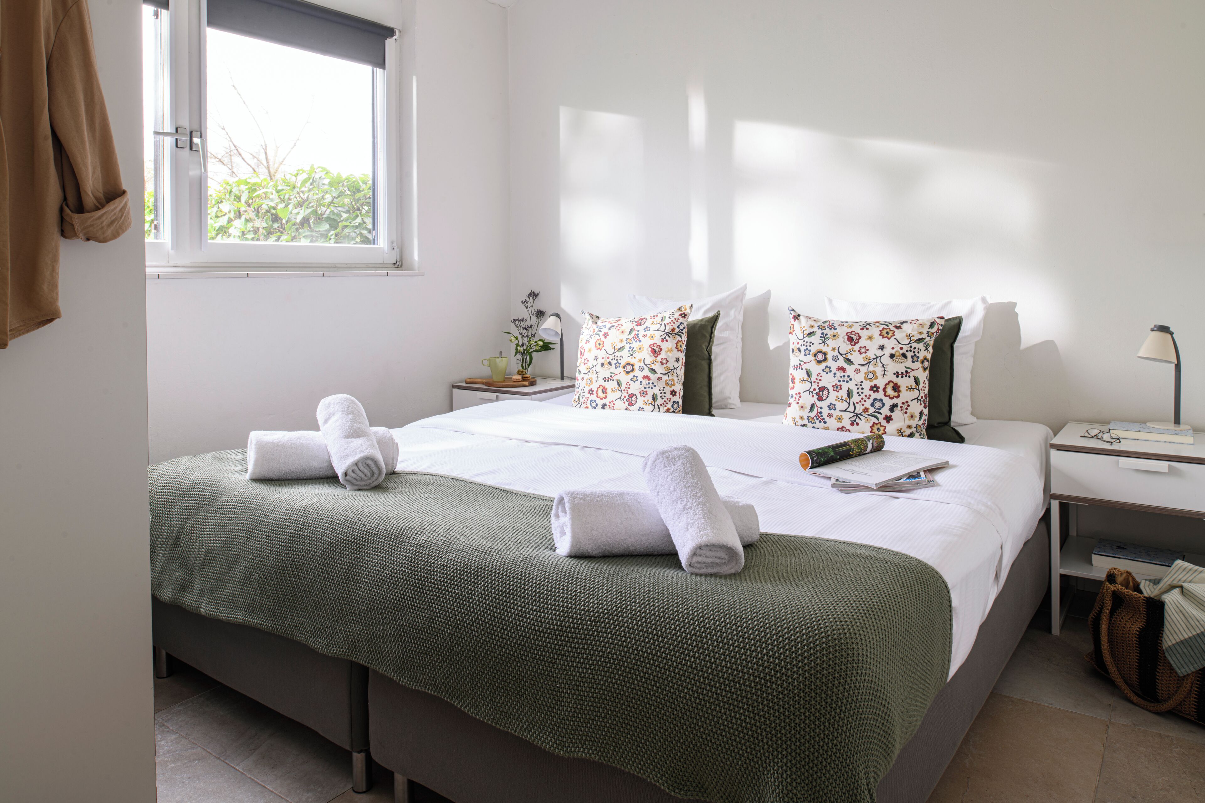 A neatly made bed with green bedding, a bedside table with a lamp, and a window with a view of greenery.