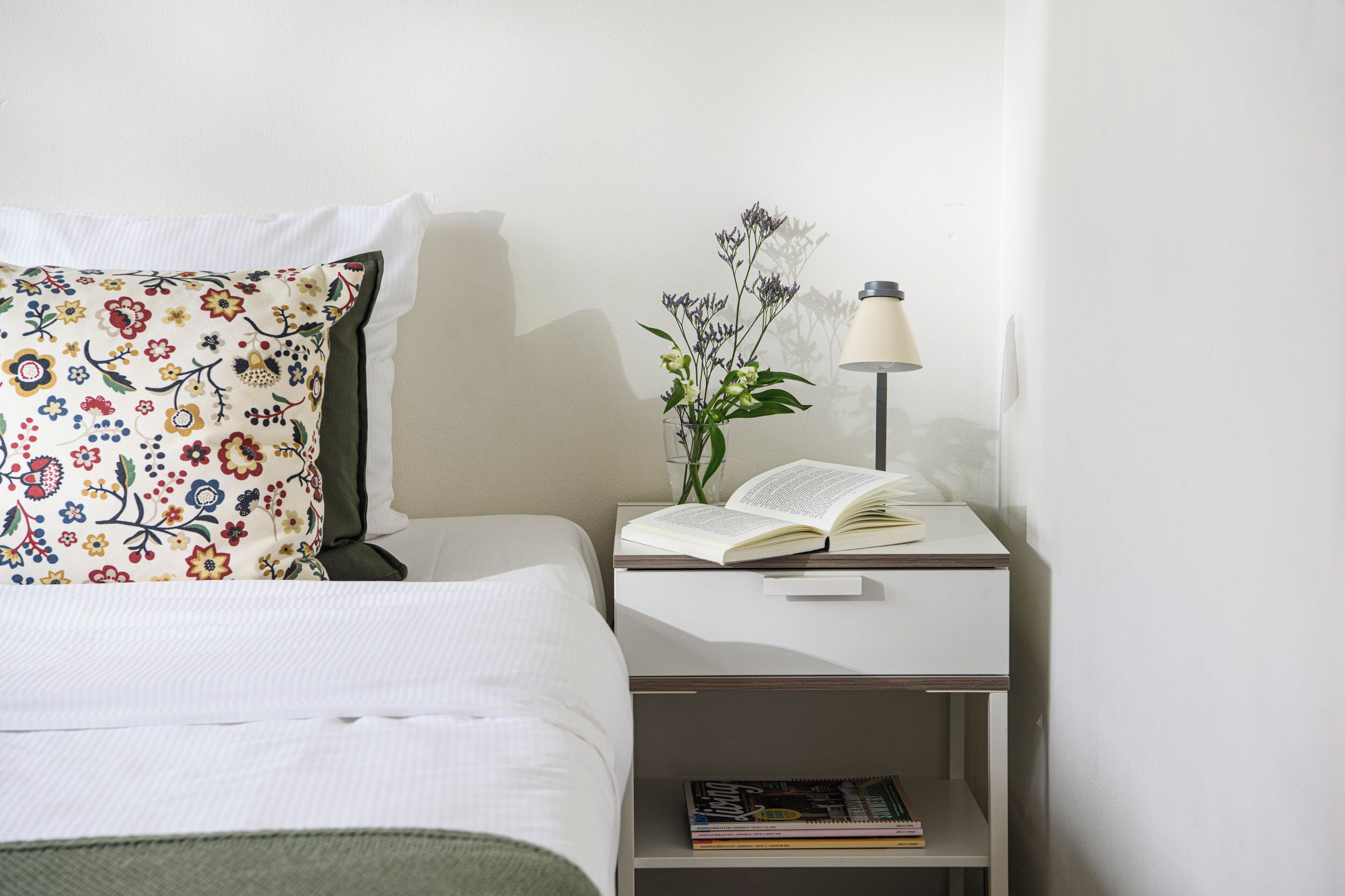 Una cama bien tendida con un cojín floral, una mesita de noche con una lámpara, y un pequeño estante con libros.