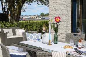 An outdoor seating area with wicker furniture, a table with a vase of flowers, a plate of cookies, and a bottle of wine. - Loosdrecht Rien van den Broeke Village (Loosdrecht)