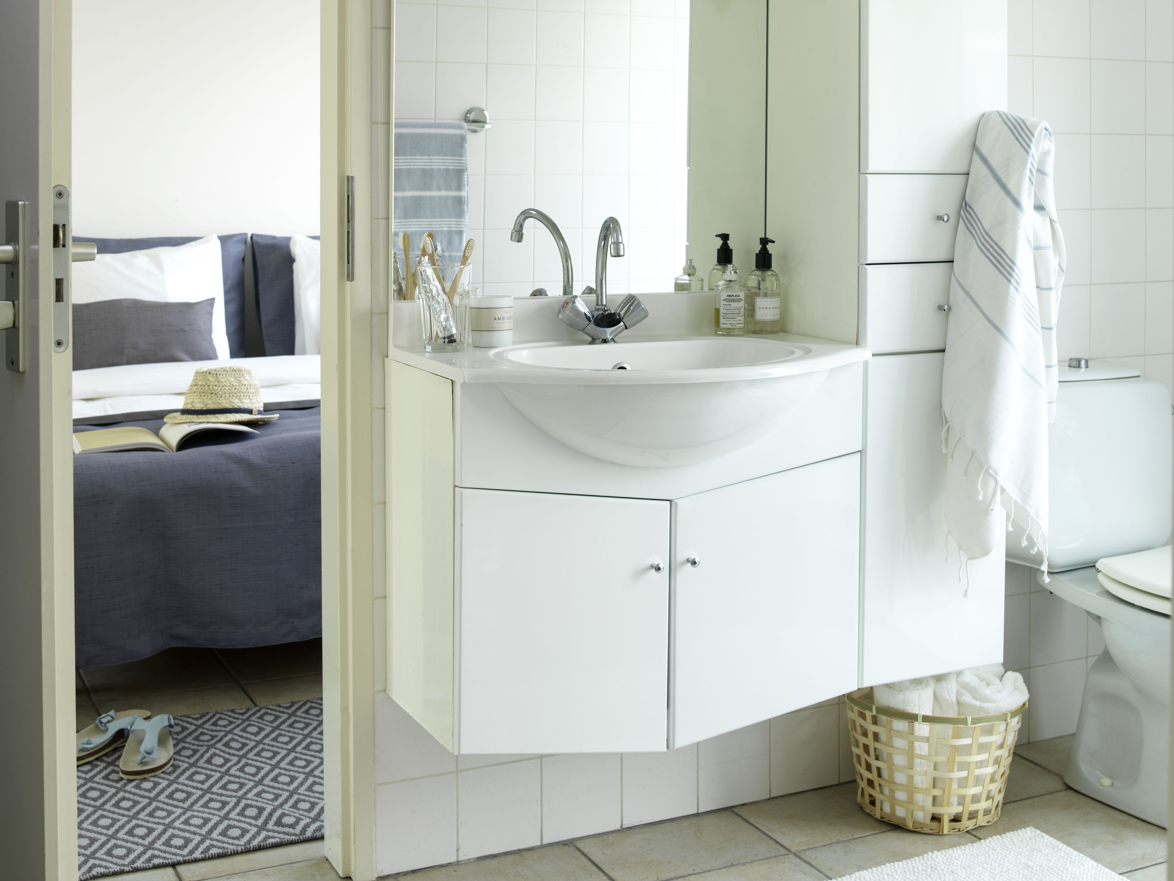 Een moderne badkamer met een witte wastafel, spiegel en toilet. Er staat een nachtkastje met een boek en een hoed.
