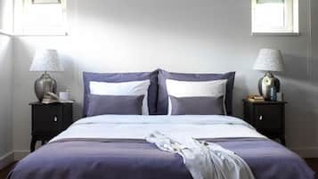 Una cama bien hecha con colcha morada, dos mesitas de noche con lámparas y una ventana con persianas.