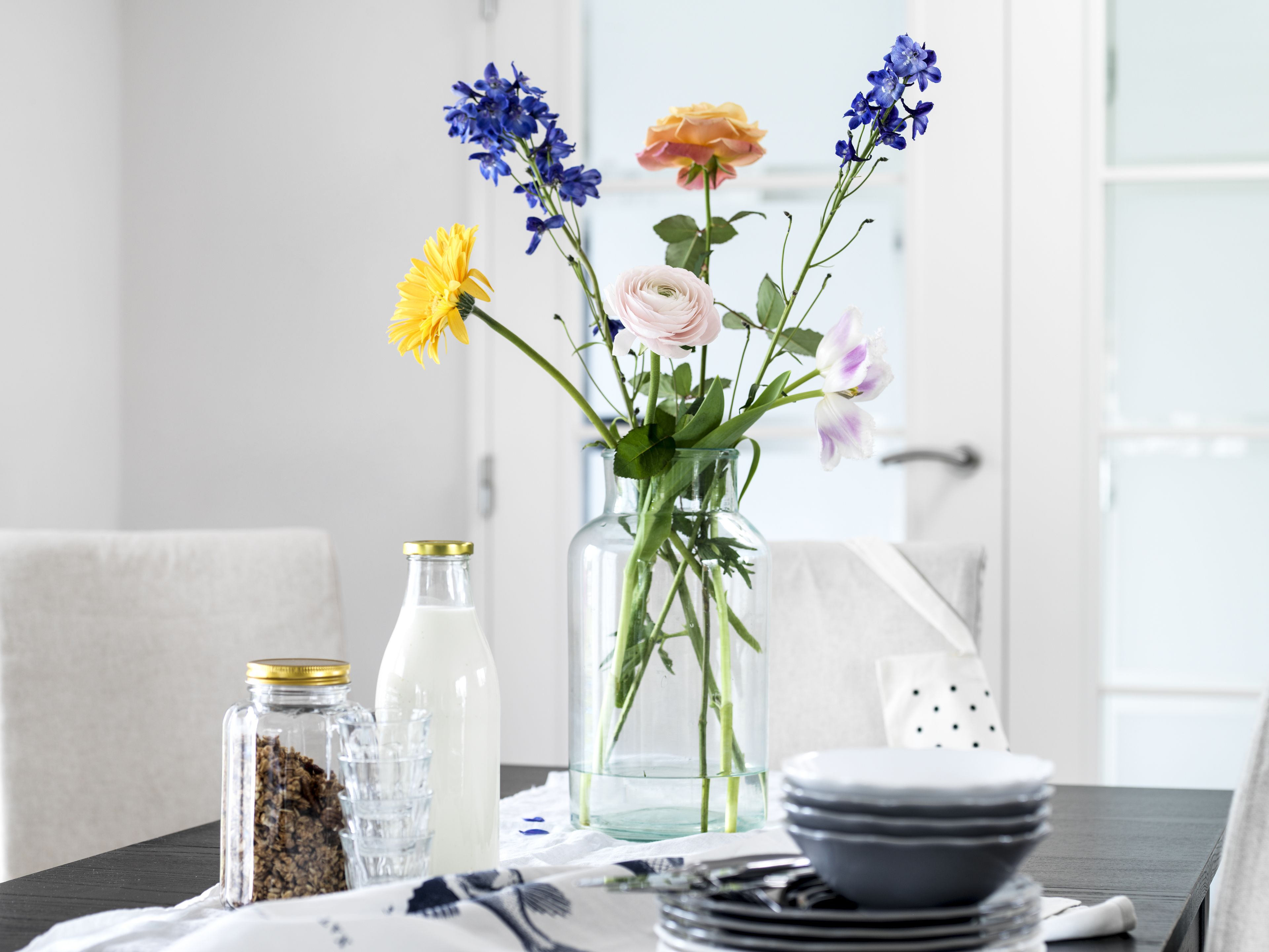 Una mesa de comedor con un florero, una botella de leche y un tarro de granola.