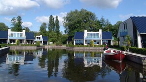 A row of modern houses with a calm water body in the foreground, a red boat docked on the right, and a clear blue sky.