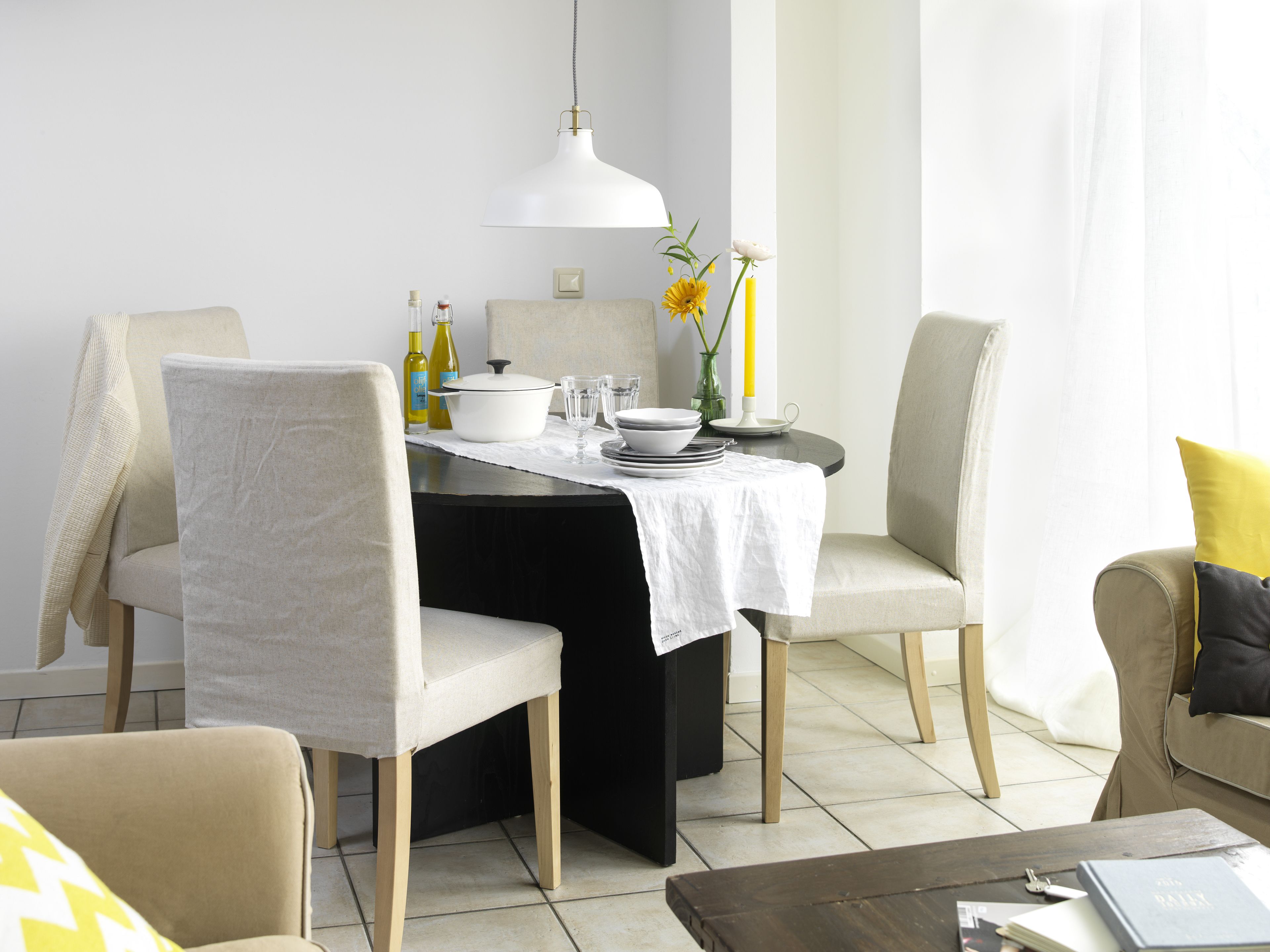 Un comedor con una mesa dispuesta para cuatro, sillas, mantel blanco, lámpara colgante blanca y un jarrón con flores amarillas.